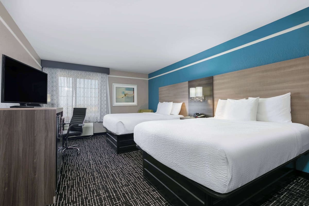 A hotel room with two beds, a desk with a TV, and a large window with curtains.