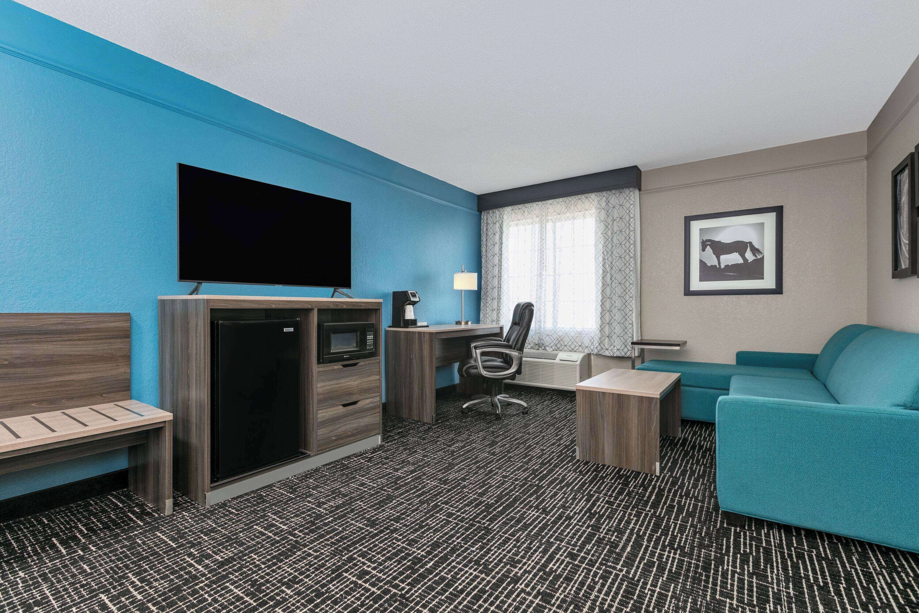 Habitación de hotel con televisor de pantalla plana, escritorio con computadora, sofá color turquesa, pequeña mesa de centro y banco.