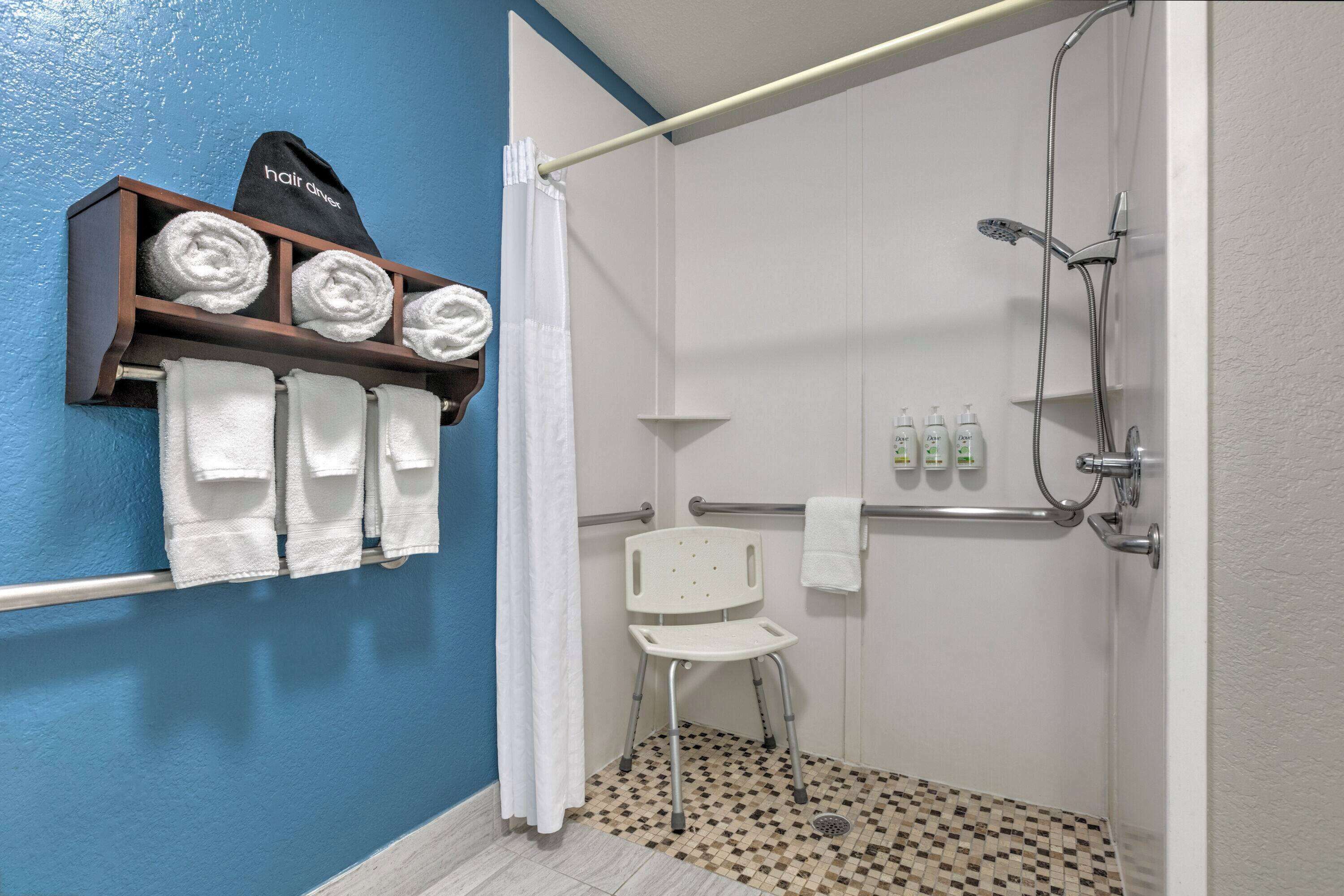 A bathroom with a shower, a white chair, a towel rack with towels, and a shelf with folded towels.