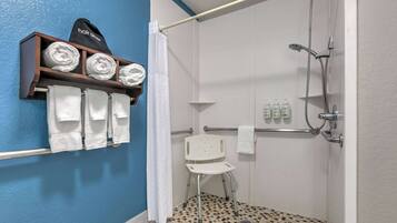 A bathroom with a shower, a white chair, a towel rack with towels, and a shelf with folded towels.