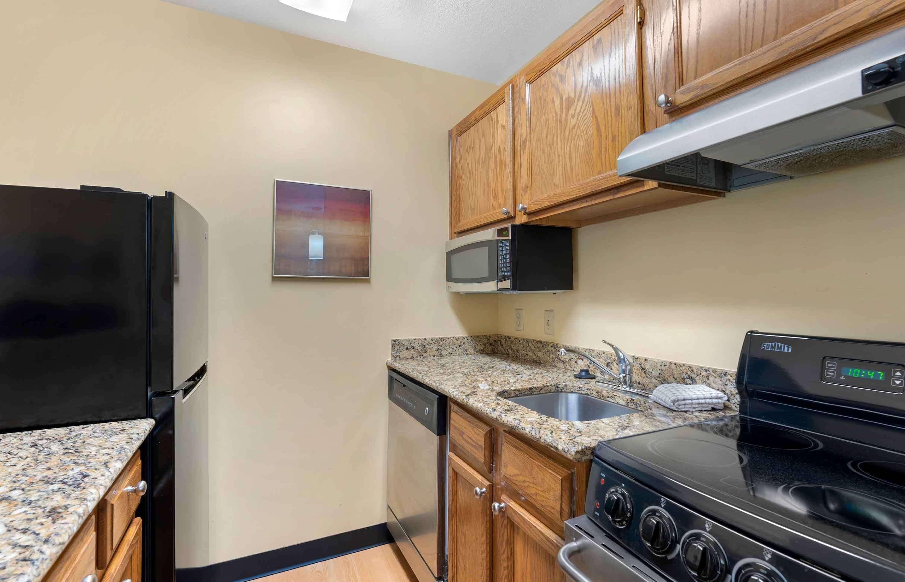 A kitchen with a black refrigerator, a microwave, a dishwasher, a granite countertop, a stainless steel sink, wooden cabinets, and a black stove.