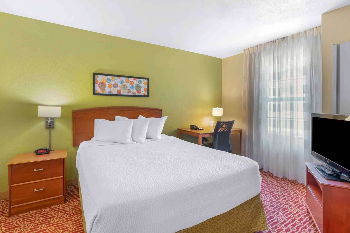 A hotel room with a bed, bedside tables, a desk, a chair, a television, and a painting on the wall.
