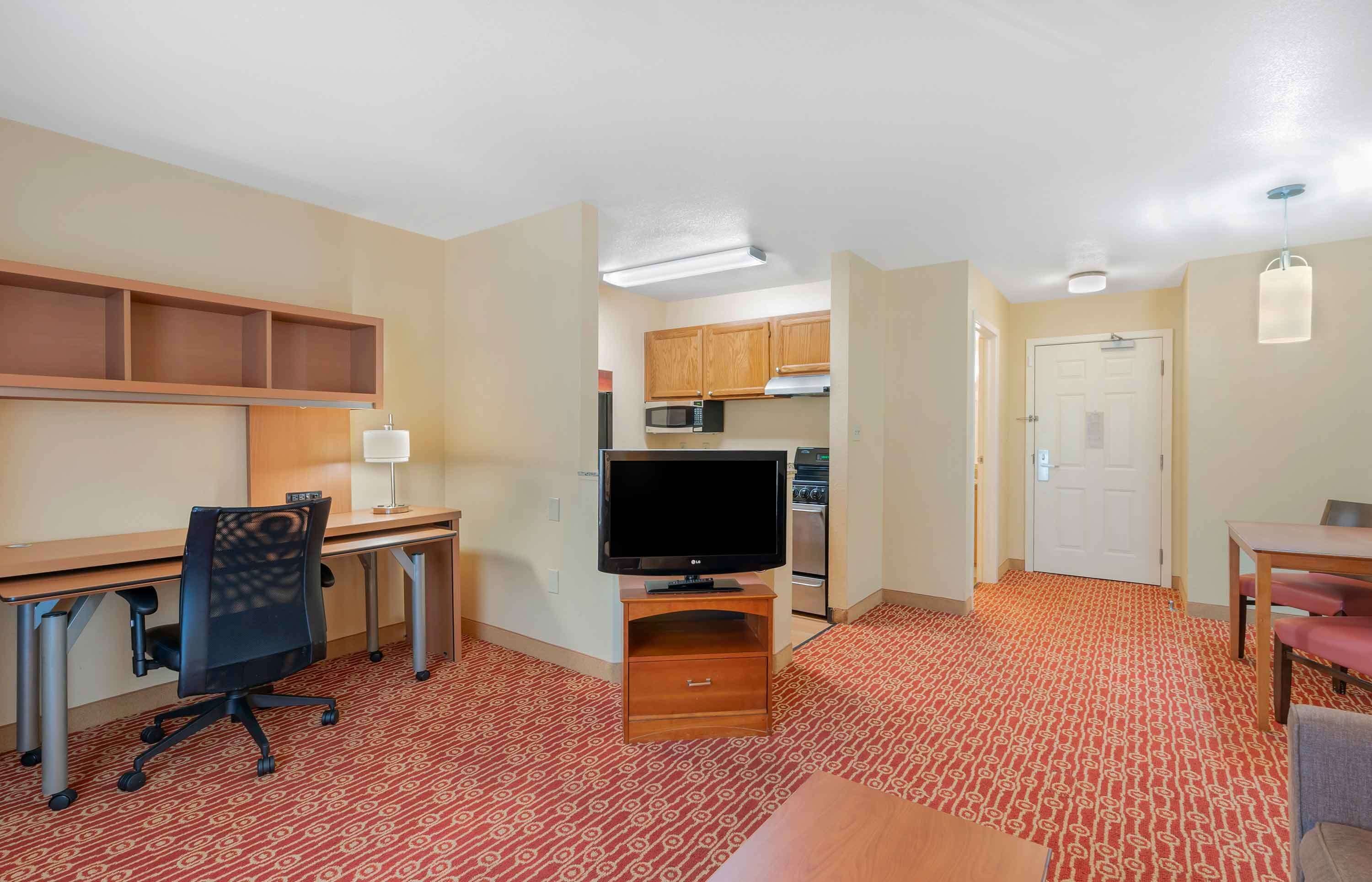 A hotel room with a kitchenette, a desk with a computer, a television, a dining area, and a patterned carpet.