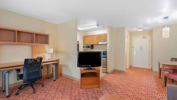 A hotel room with a kitchenette, a desk with a computer, a television, a dining area, and a patterned carpet.