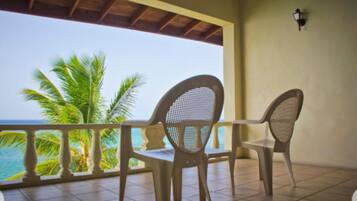 A balcony with two wicker chairs, a tiled floor, and a view of the ocean and palm trees.