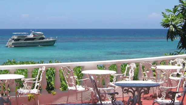 A balcony with white metal chairs and tables, overlooking a turquoise sea with a yacht.