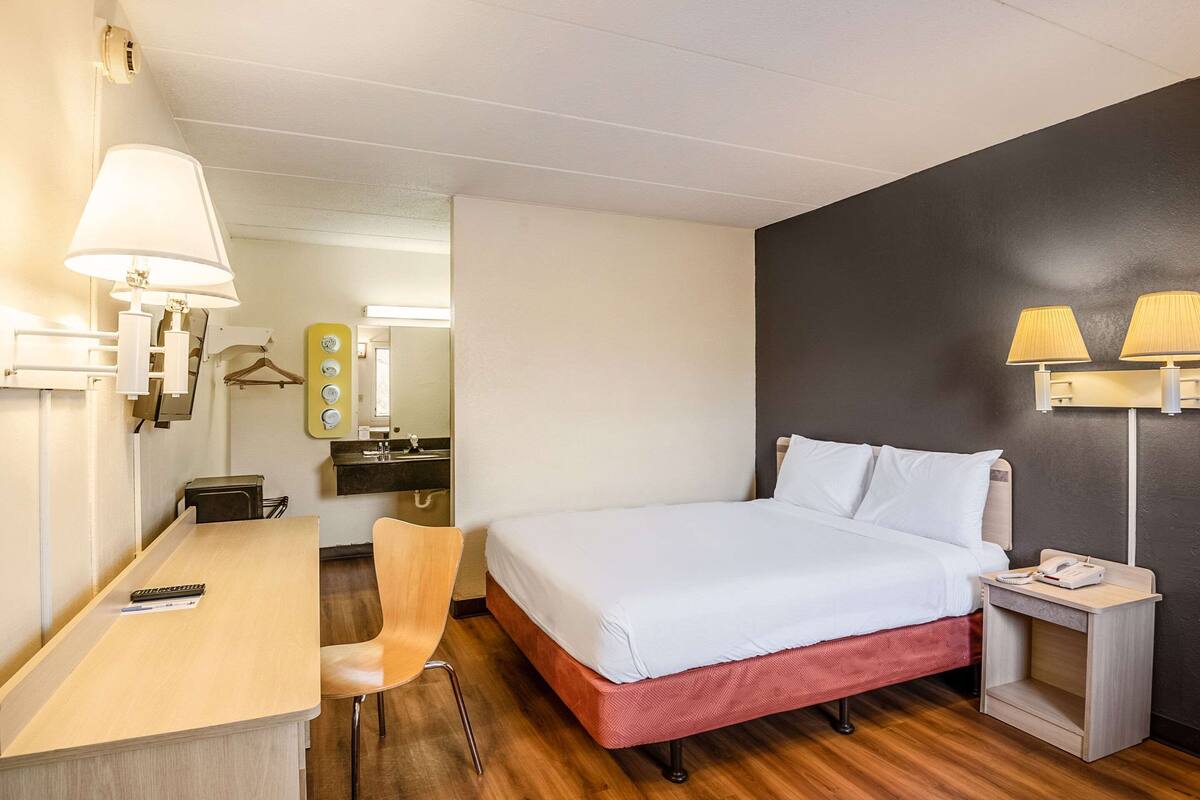 A hotel room with a bed, bedside table, chair, desk, lamp, and a small bedside table with a phone.