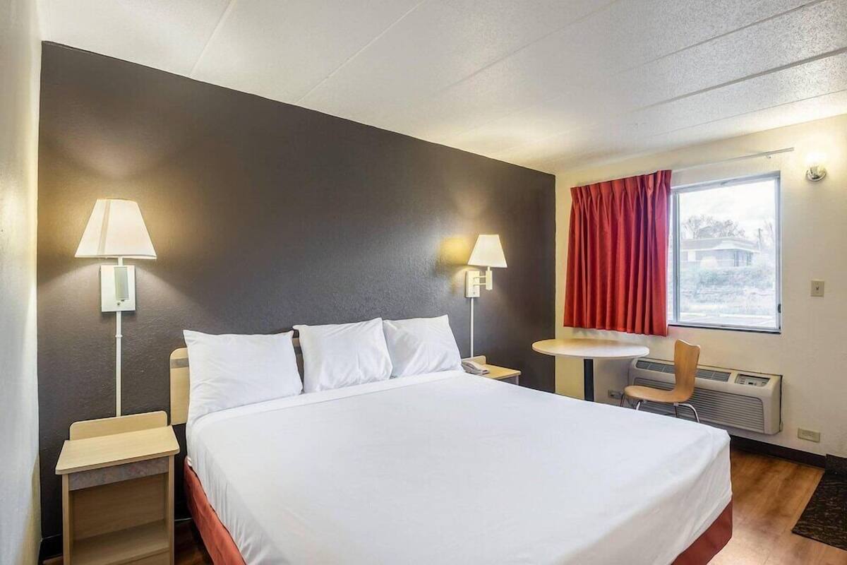 A hotel room with a bed, two bedside lamps, a small table, a chair, a window with curtains, and an air conditioning unit.