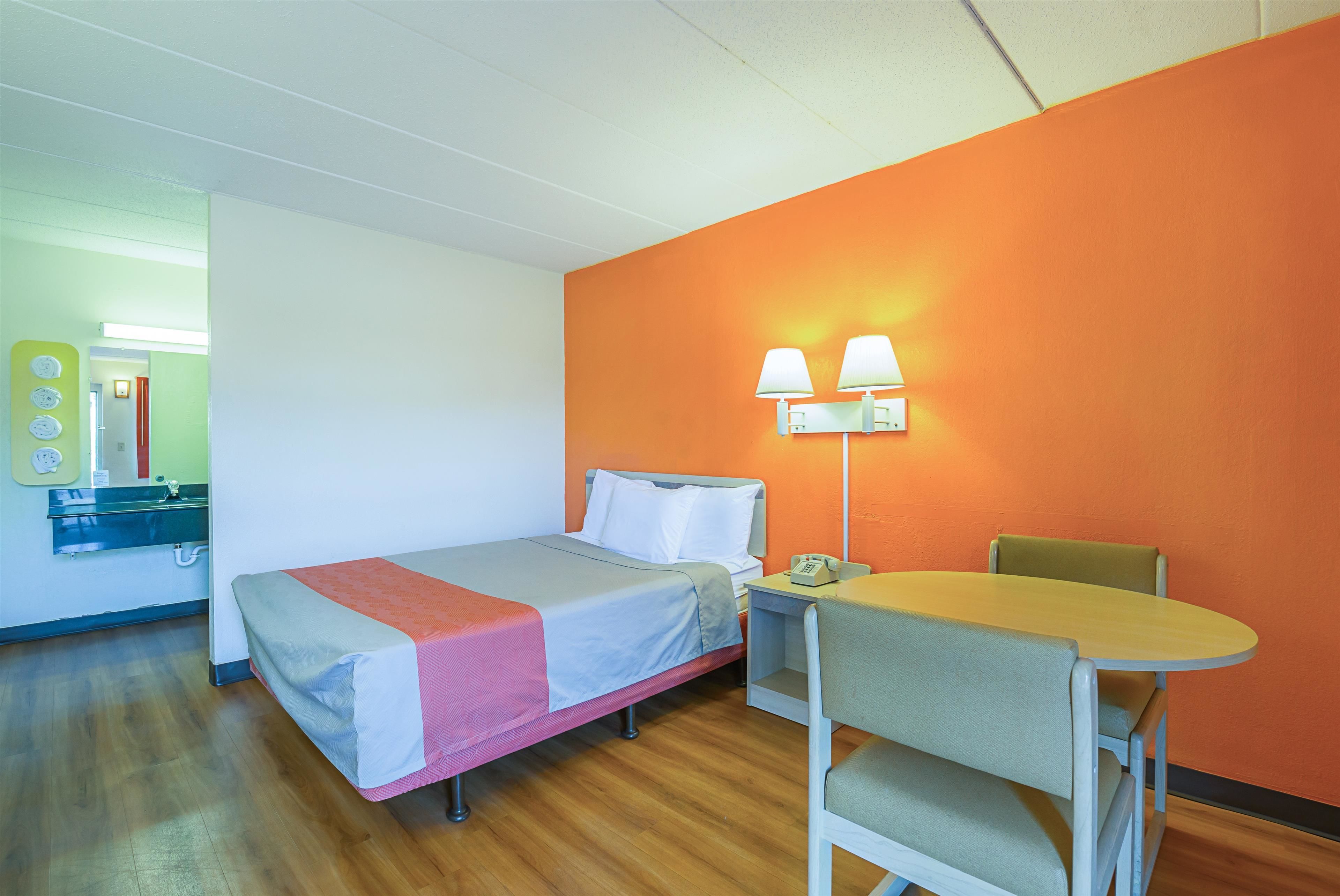 A single bed with a red blanket, a small table with a chair, and a nightstand with a lamp.