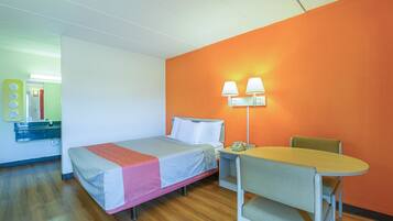 A single bed with a red blanket, a small table with a chair, and a nightstand with a lamp.