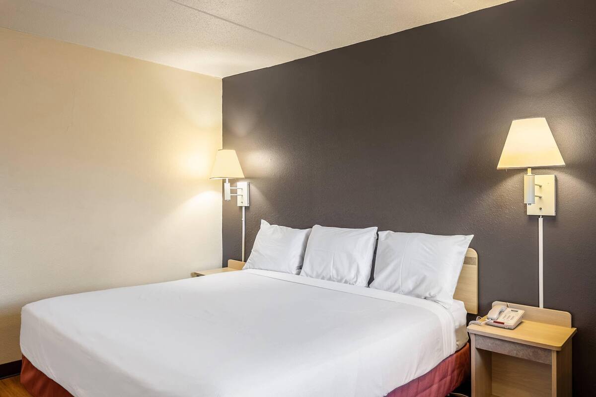 A hotel room with a large bed, two bedside lamps, a nightstand with a phone, and a wall-mounted light switch.