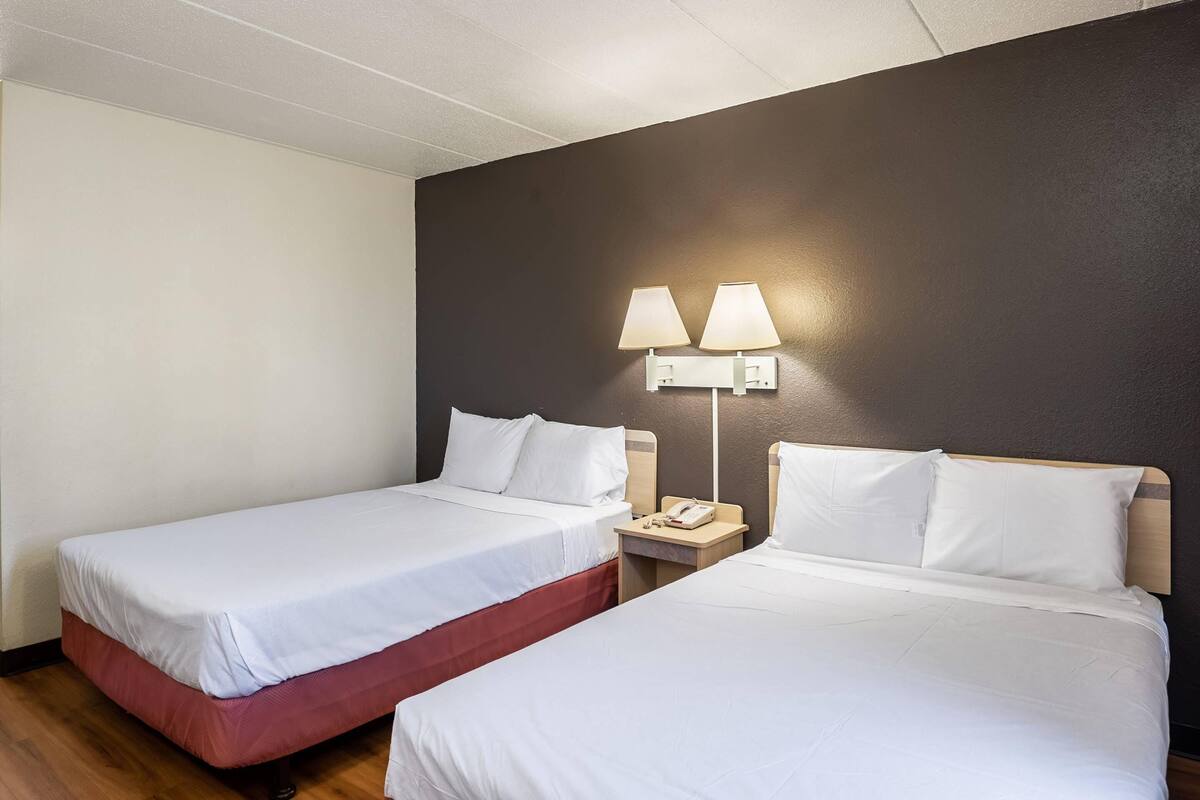 Two single beds with white linens, a wooden nightstand with a lamp, and a dark-colored wall.