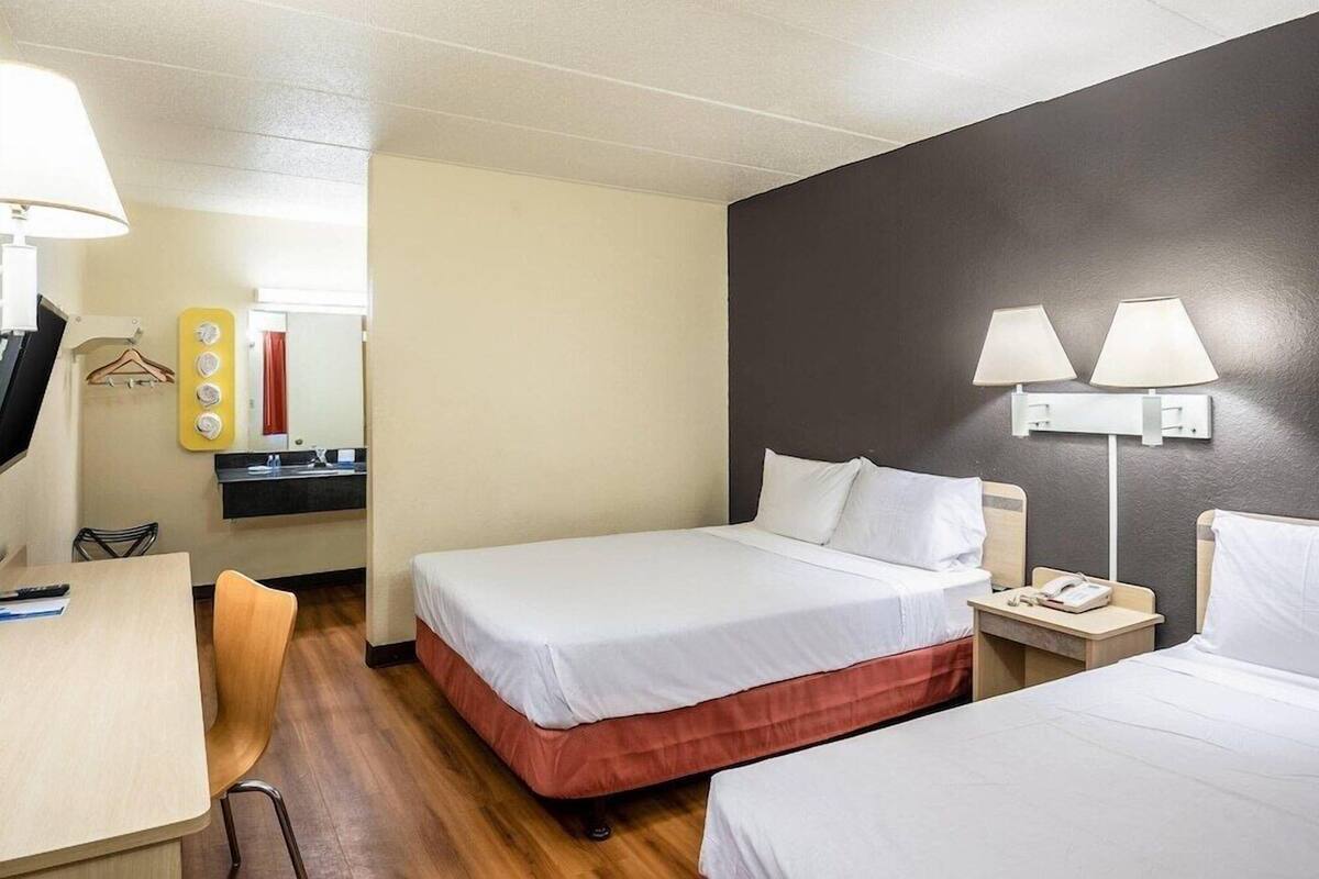 A hotel room with two beds, a desk, a chair, a small table, a mirror, and a wall-mounted lamp.