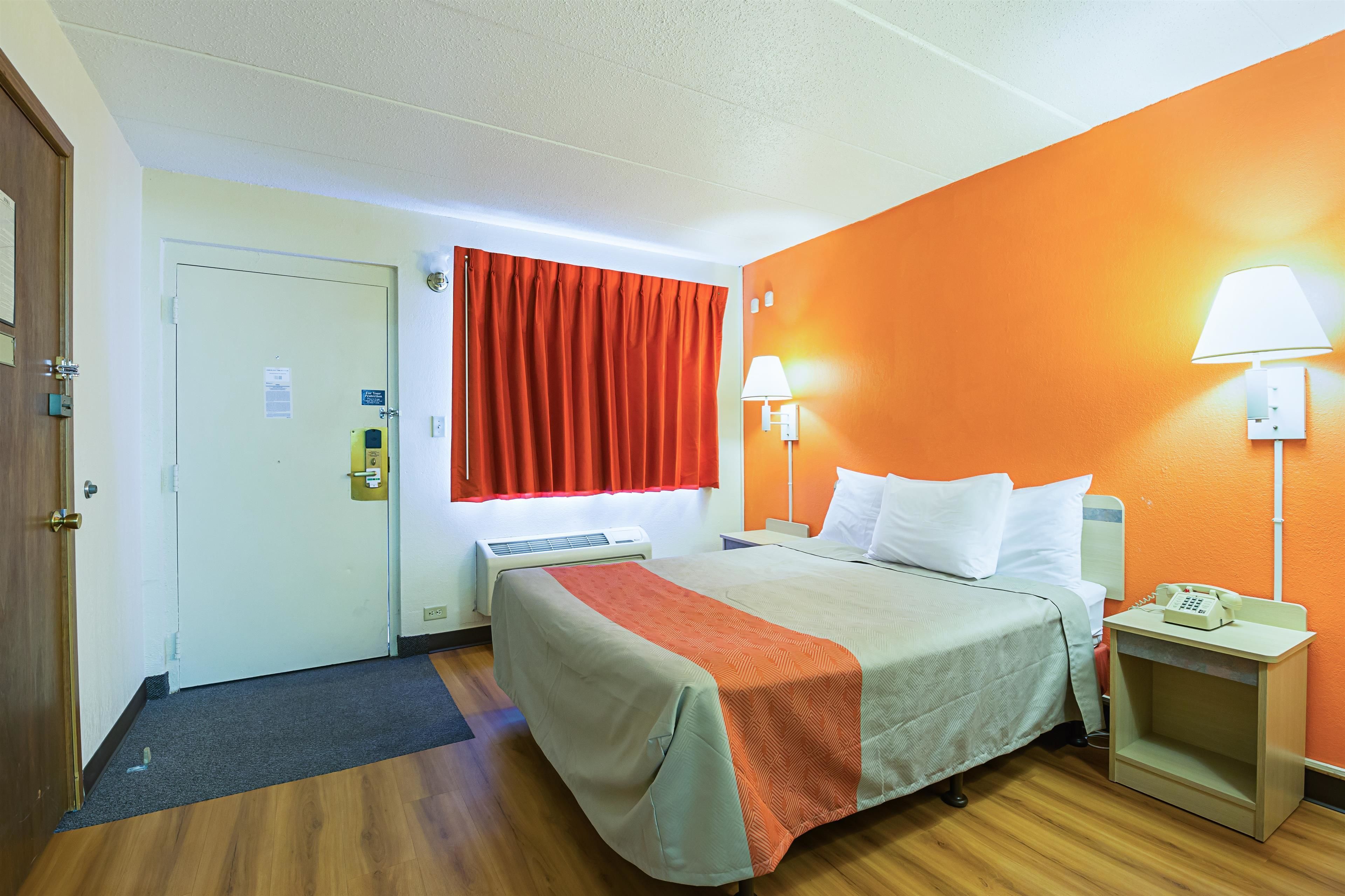A hotel room with a bed, two bedside lamps, a nightstand with a phone, a window with red curtains, and a door with a lock.