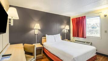 A hotel room with a bed, a desk, two chairs, a TV, and a window with red curtains.