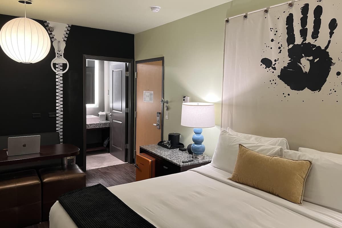 A hotel room with a bed, desk, lamp, and a wall with a handprint design.