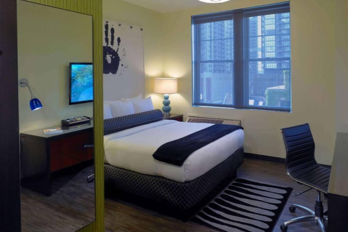 A hotel room with a bed, desk, chair, lamp, artwork, and a window with a city view.