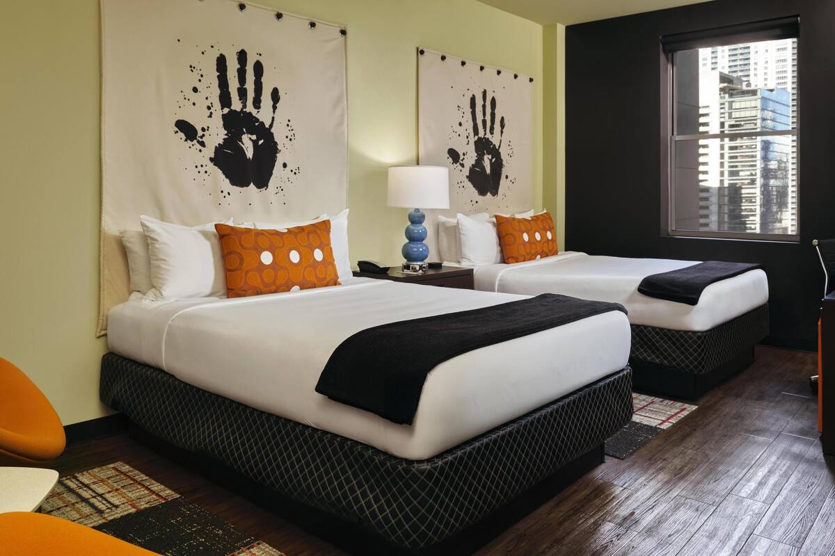 A hotel room with two beds, a nightstand with a lamp, a window with a view of buildings, and a handprint artwork on the wall.