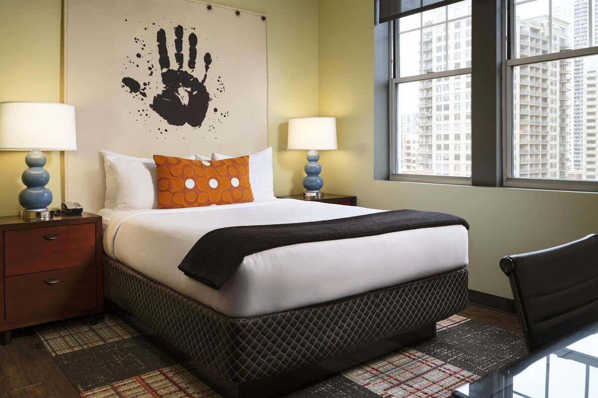 A hotel room with a bed, two bedside tables with lamps, a large window with a city view, and a handprint artwork on the wall.