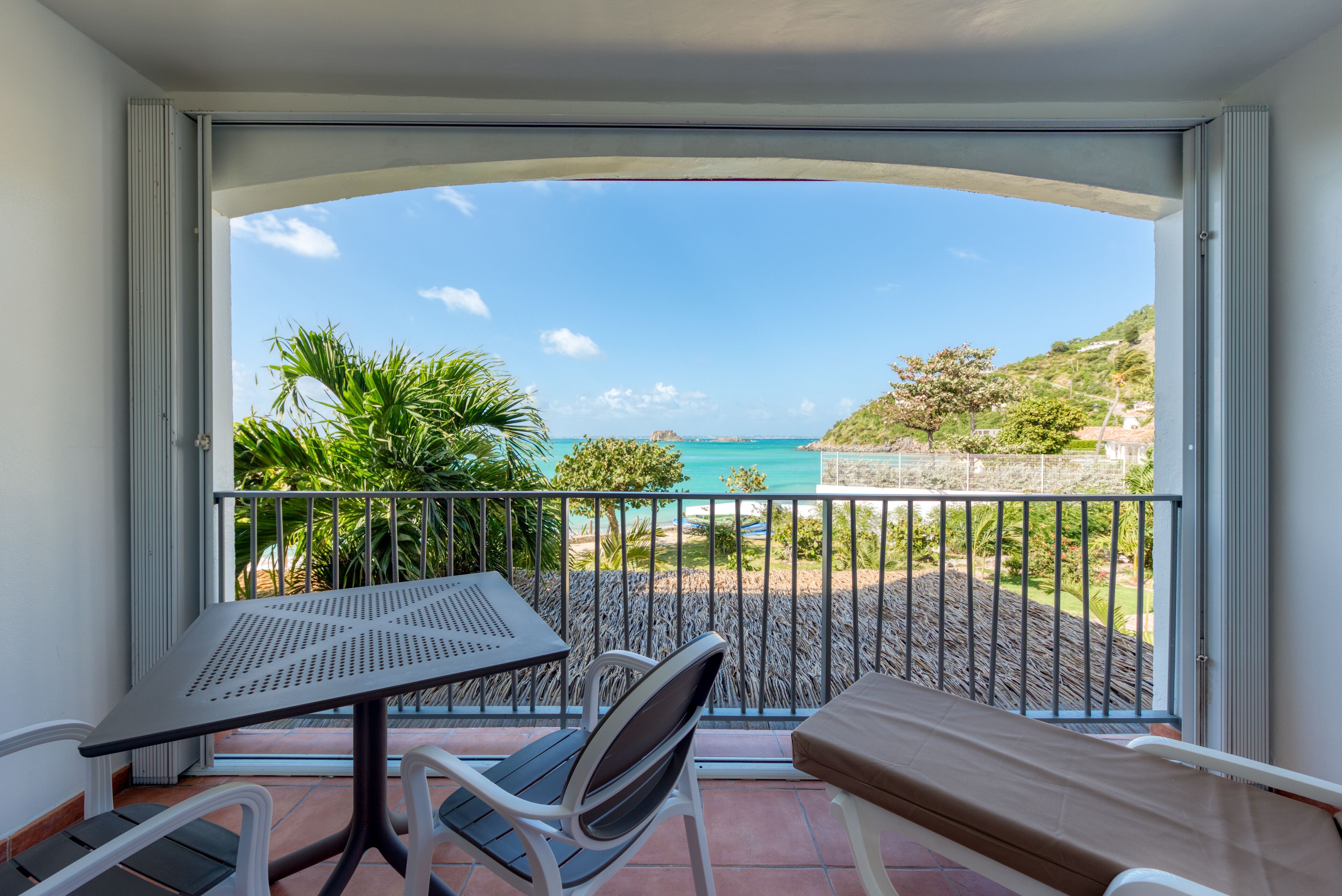 A balcony with a table and chairs, overlooking a coastal landscape with trees and a view of the sea.