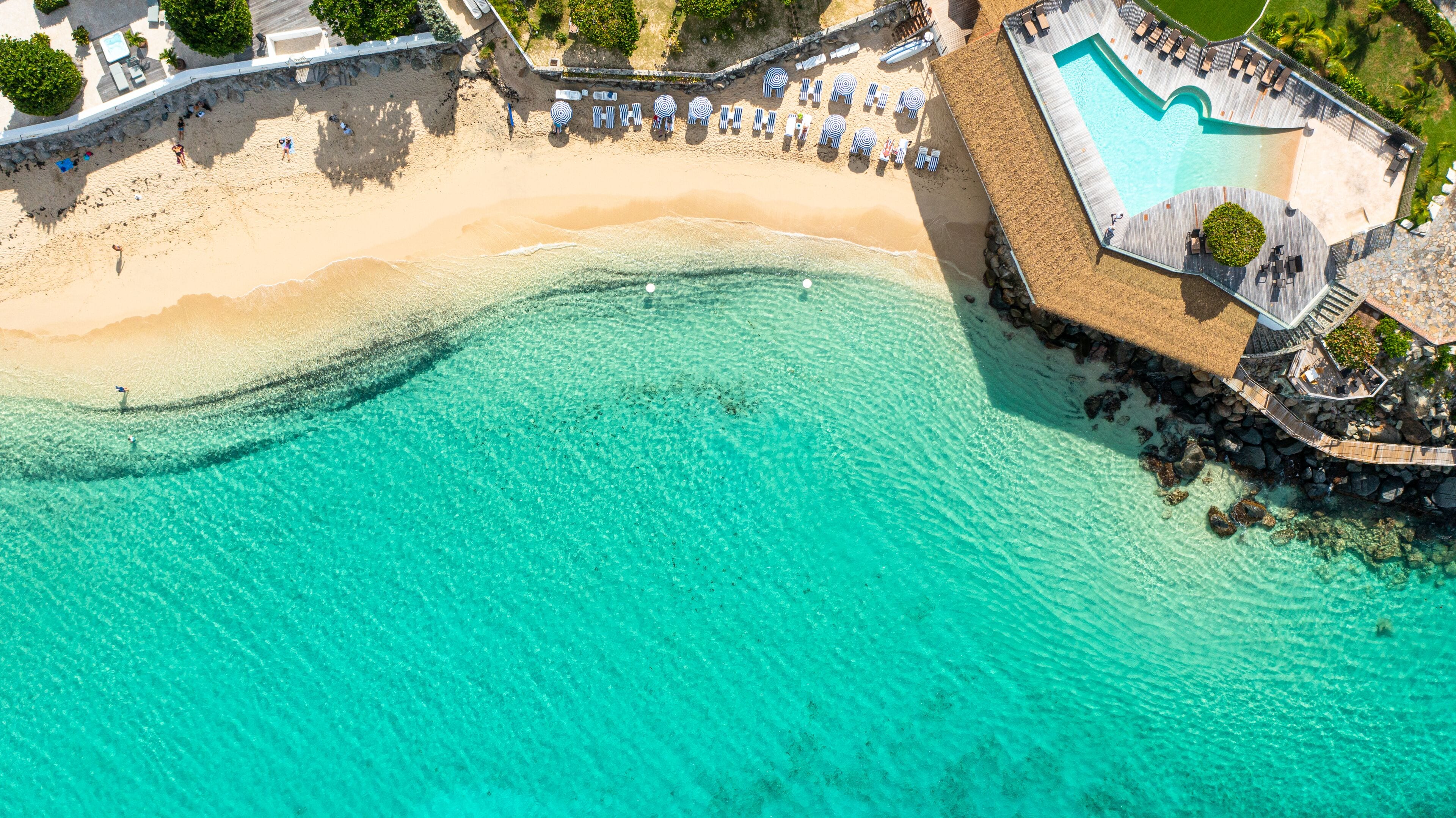 An aerial view of a beach resort with a clear turquoise pool, a sandy beach, and a wooden deck with lounge chairs.