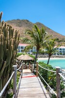 Un muelle de madera que conduce a una playa con una cabaña de techo de paja, rodeada de cactus y palmeras, con una montaña al fondo.