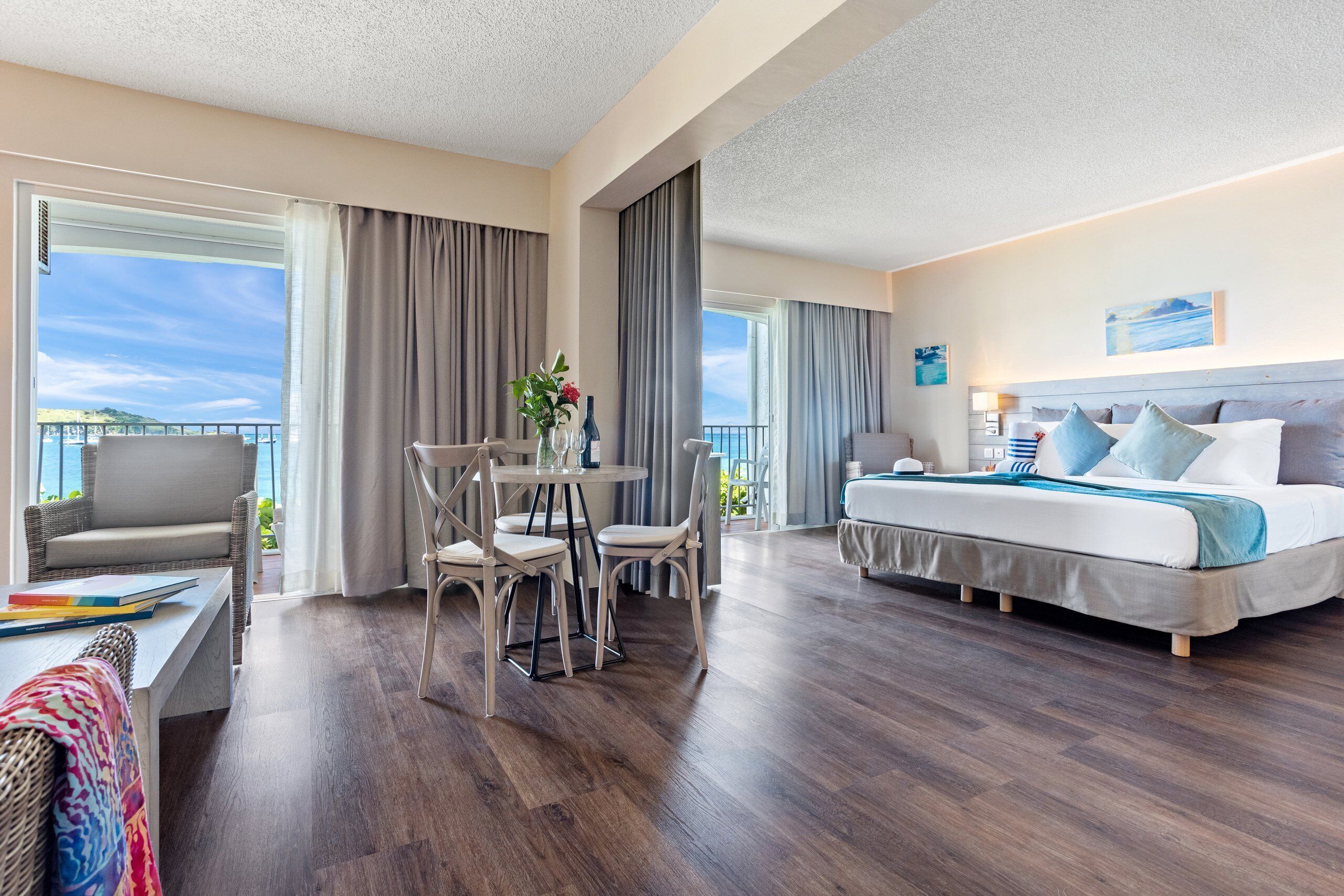 A hotel room with a large bed, a dining table with chairs, a balcony with curtains, and a view of the sea.