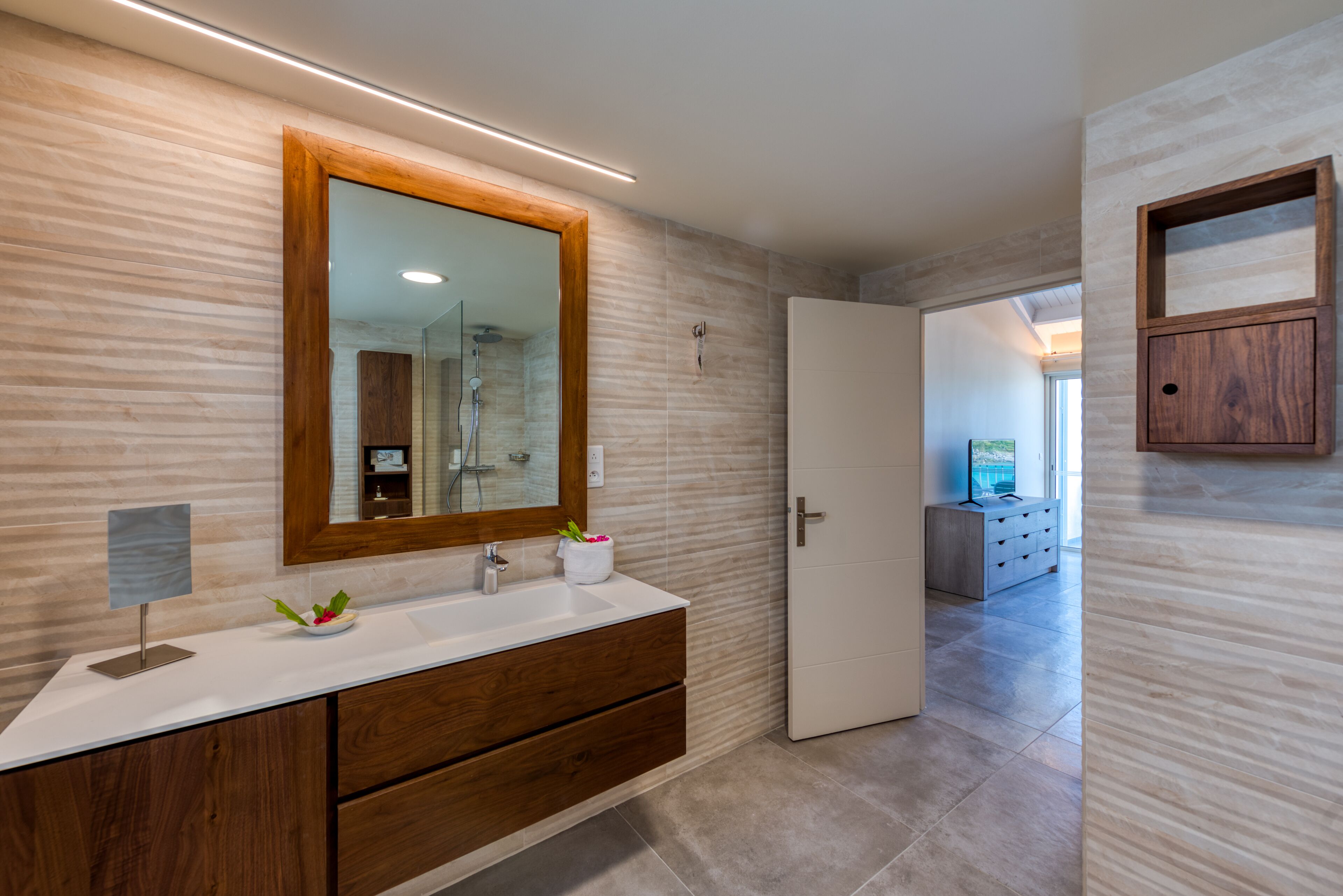 Un baño moderno con un mueble de tocador de madera, un espejo grande y una zona de ducha visible al fondo.