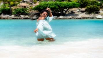 A person in a white dress standing in shallow water at a beach with clear blue water and palm trees in the background.