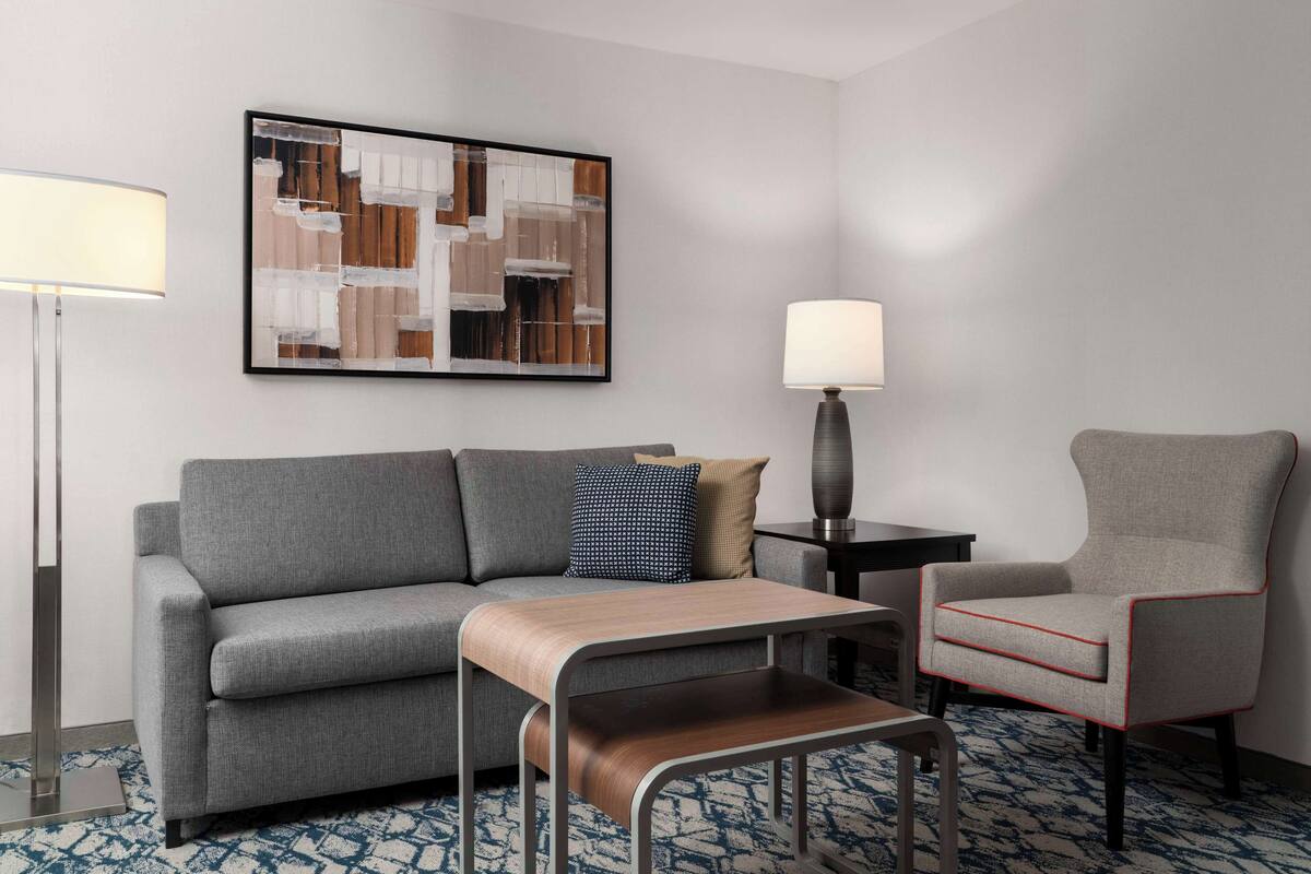 Een moderne hotelkamer met een grijze bank, een houten bijzettafeltje, een grijze fauteuil en een lijst met een abstract schilderij aan de muur.