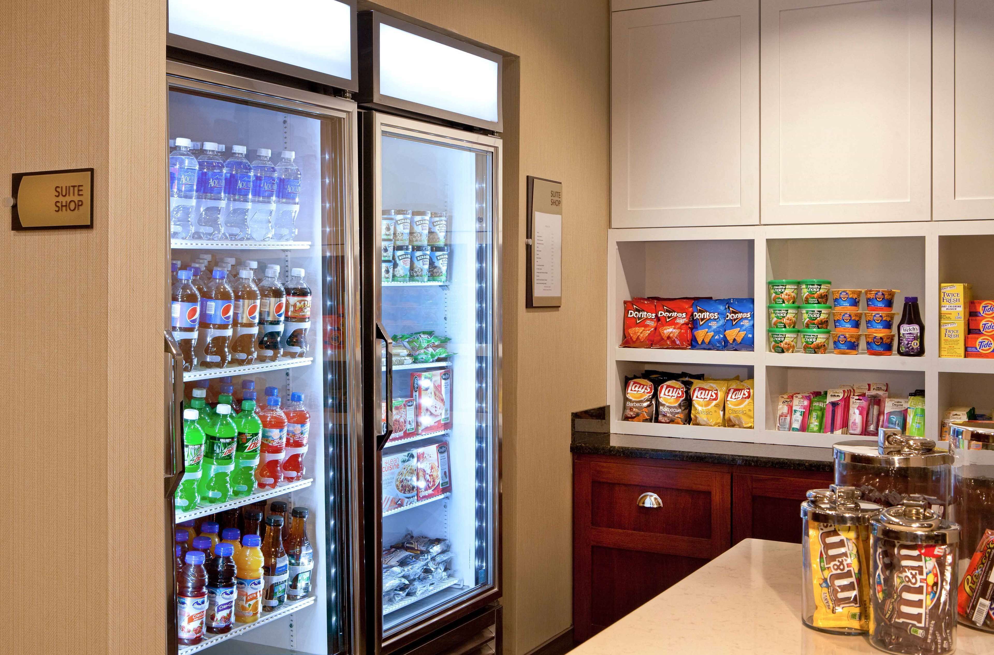 A hotel suite with a mini-fridge stocked with beverages and snacks, a minibar with various food items, and a small kitchen area with a table and cabinets.