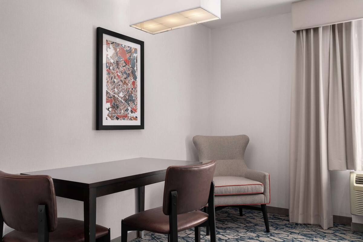 Een hotelkamer met een eettafel, twee stoelen, een lijst met abstracte kunst, een raam met gordijnen en een elektrische kachel.