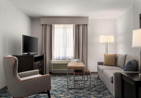 A hotel room with a flat-screen TV, a sofa, a chair, a small table, and a lamp.