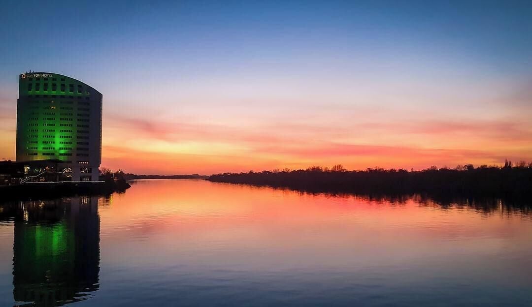 Un hôtel au design moderne, situé au bord d’une étendue d’eau paisible, dont les eaux reflètent le ciel au coucher du soleil.