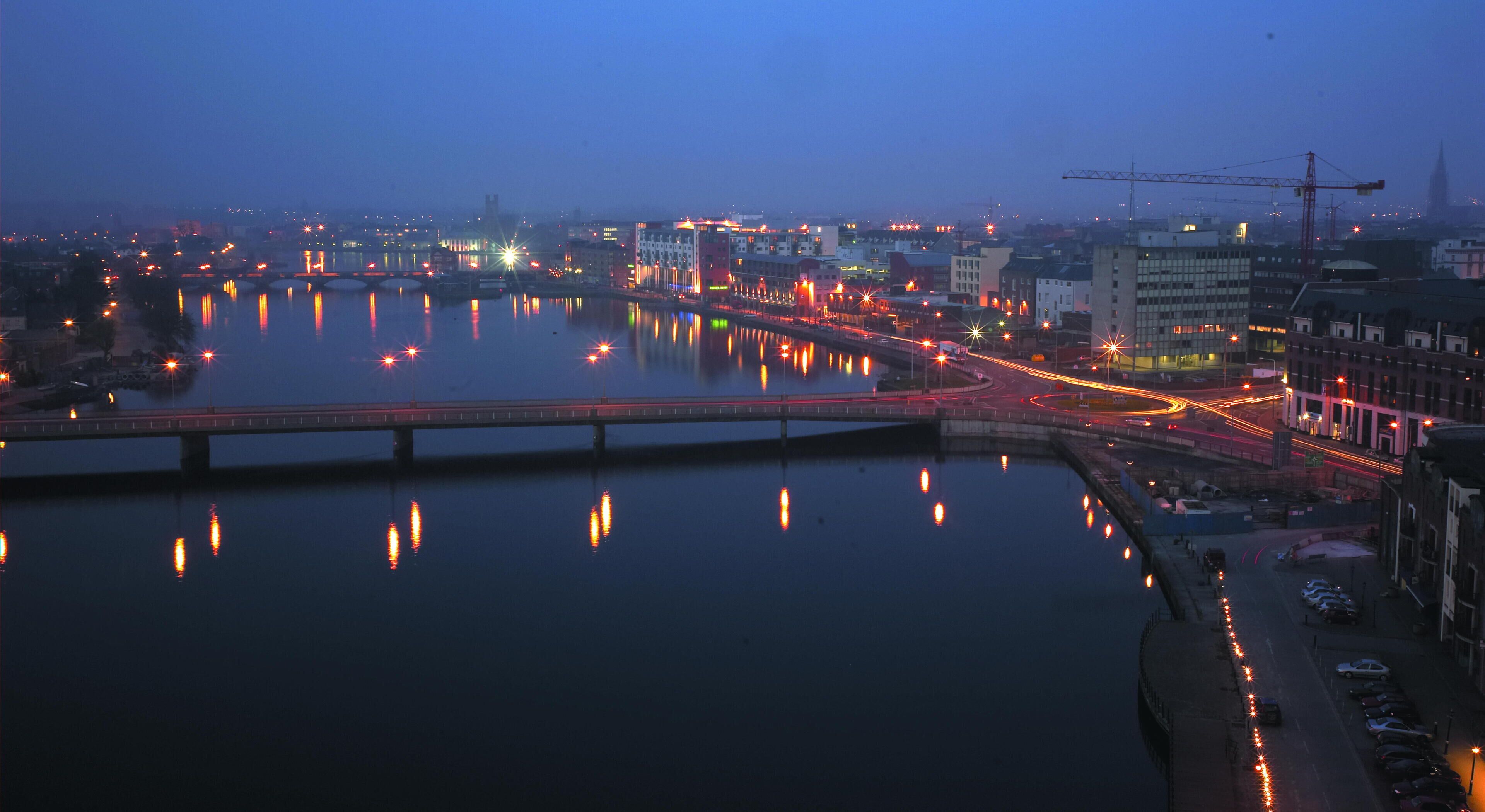 A nighttime cityscape with a river reflecting lights, a bridge, and buildings along the riverbank.