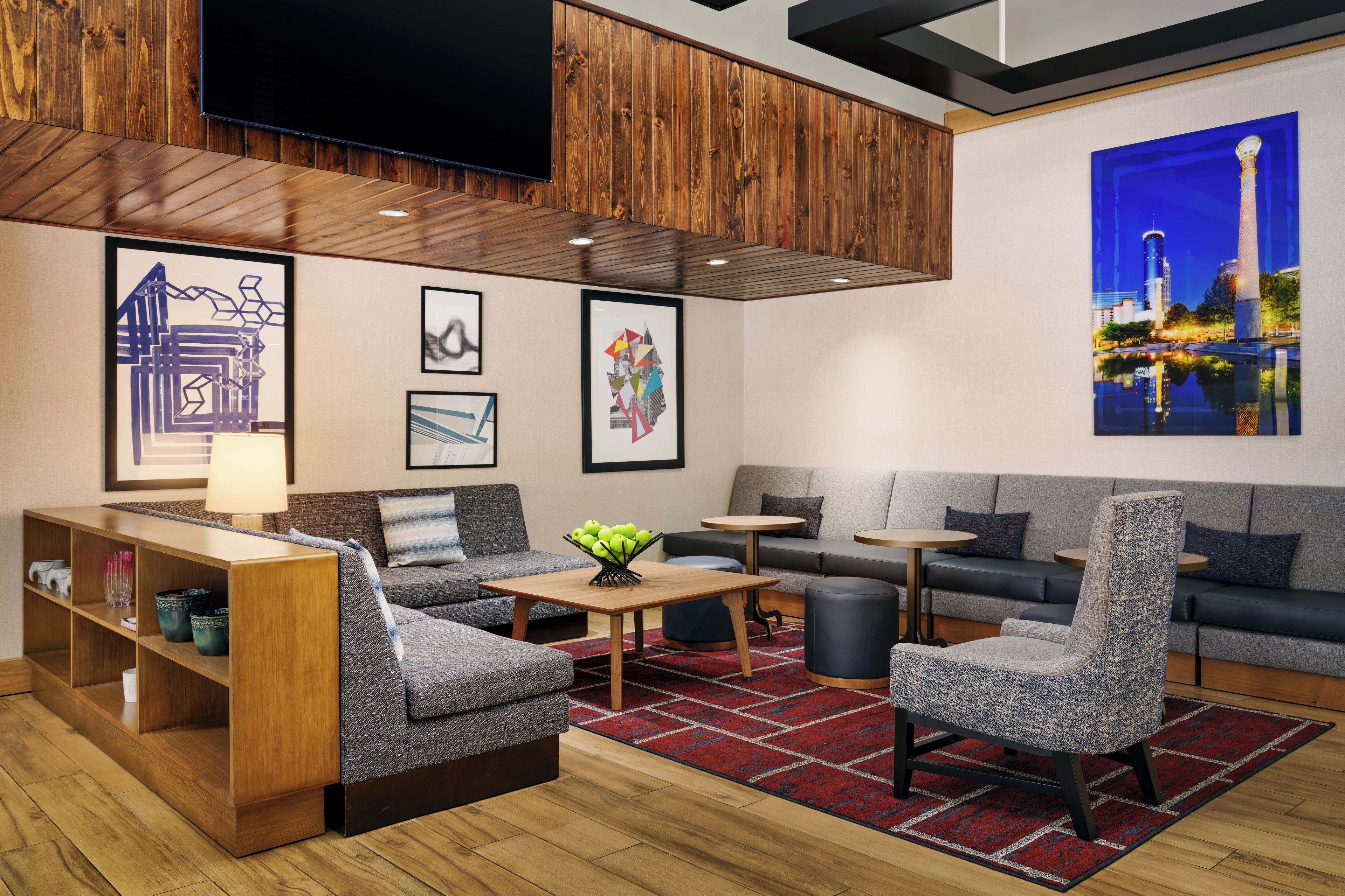 A modern hotel lobby with a wooden ceiling, a flat-screen TV, a sofa, armchairs, a coffee table, and a bookshelf.