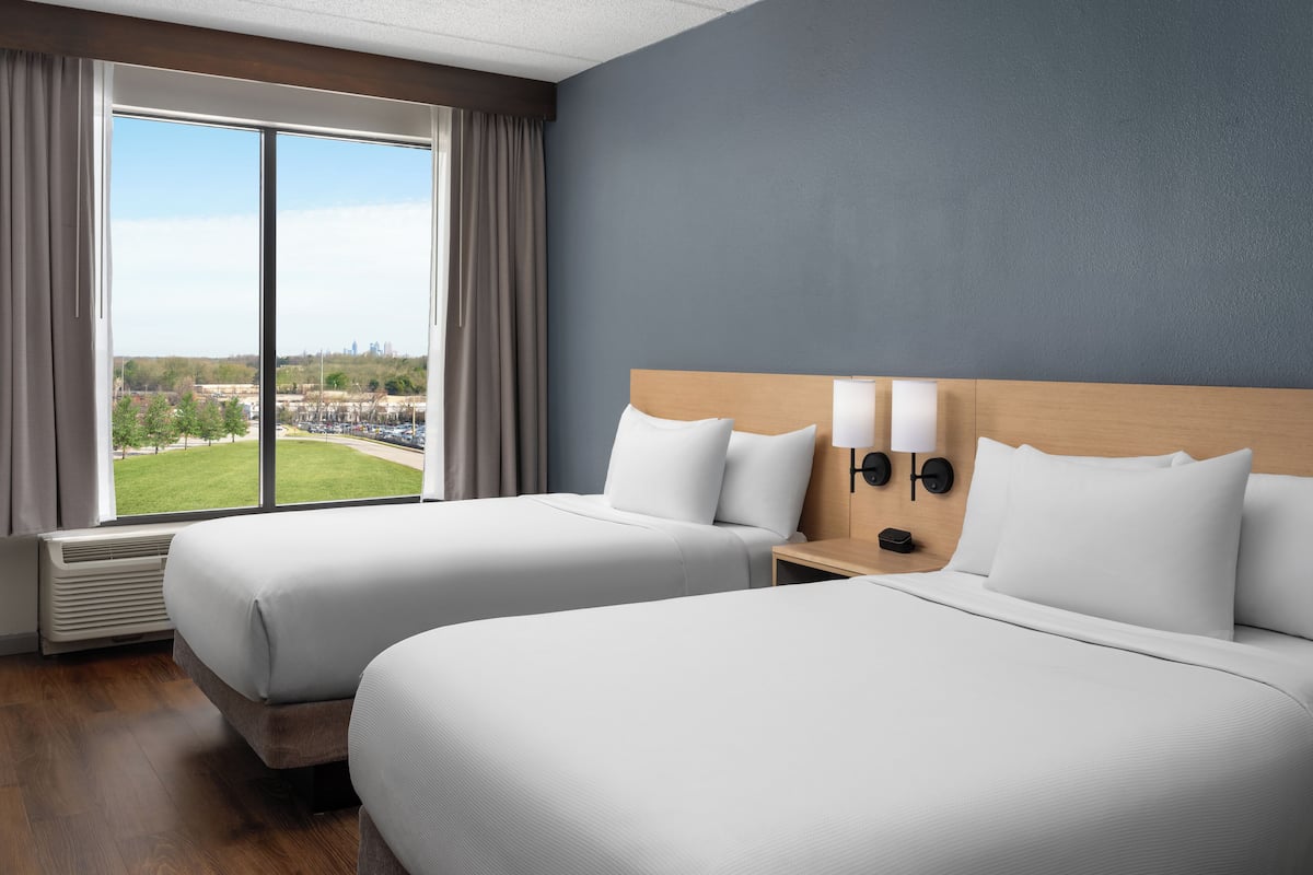 Ein Hotelzimmer mit zwei Betten, einem großen Fenster mit Vorhängen, Blick auf eine Stadtlandschaft und einer Wandlampe.
