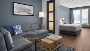 Ein modernes Hotelzimmer mit einem grauen Sofa, einem kleinen runden Tisch, einer hölzernen Bank, einem großen, gerahmten abstrakten Kunstwerk, einem Bett mit grauem Topper, einem Nachttisch mit Lampe und einem großen Fenster mit Vorhängen.