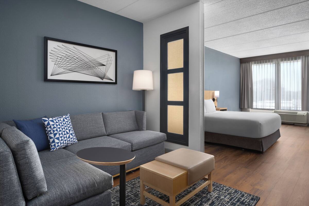 Ein modernes Hotelzimmer mit einem grauen Sofa, einem kleinen runden Tisch, einer hölzernen Bank, einem großen, gerahmten abstrakten Kunstwerk, einem Bett mit grauem Topper, einem Nachttisch mit Lampe und einem großen Fenster mit Vorhängen.