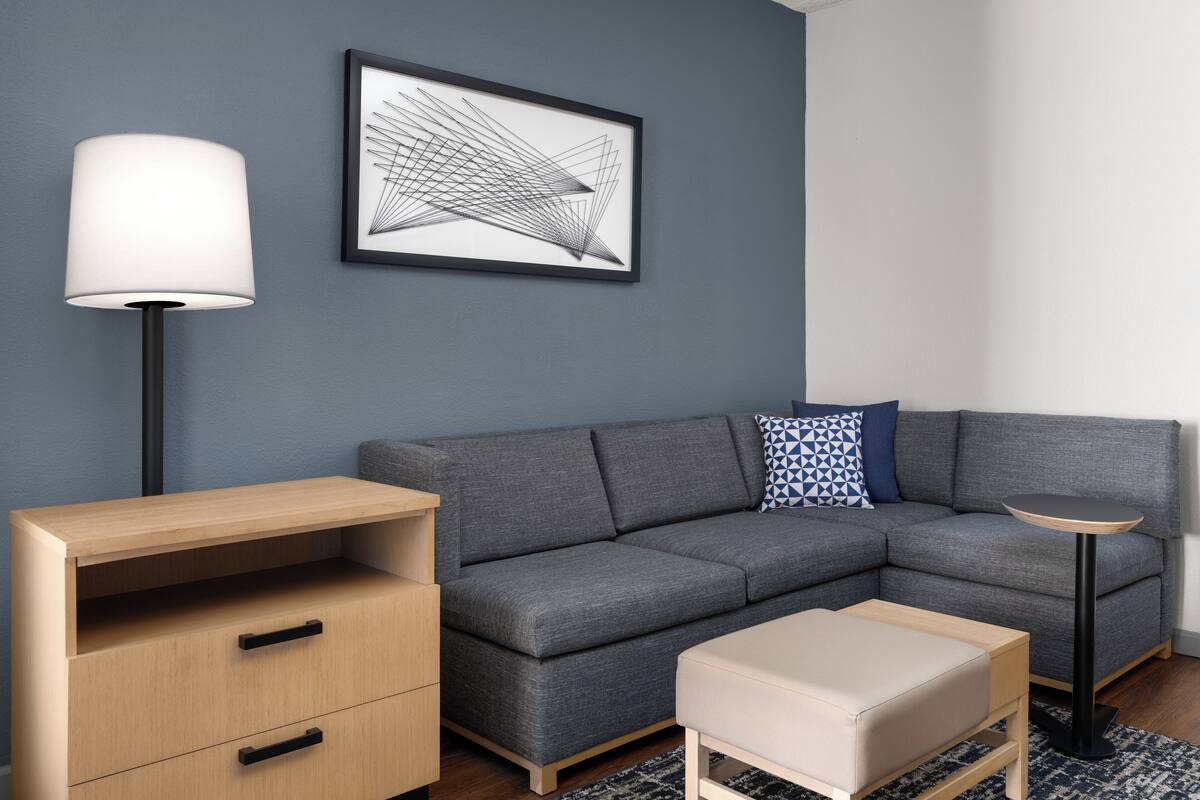 Ein modernes Wohnzimmer mit einem grauen Ecksofa, einem kleinen runden Tisch, einer Lampe und einer Holzkommode.