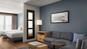 Ein modernes Hotelzimmer mit grauem Sofa, einem kleinen runden Tisch, einem hölzernen Nachttisch, einem Bett mit weißer Bettwäsche und einem großen abstrakten Gemälde an der Wand.