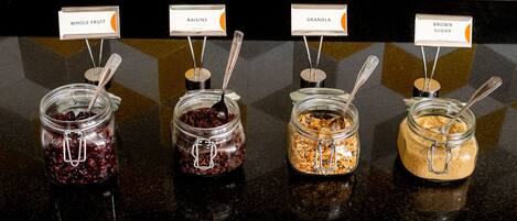 Cuatro frascos de vidrio con etiquetas sobre una encimera negra, que contienen frutas secas, granola y azúcar moreno.