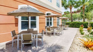 An outdoor patio with tables and chairs, surrounded by palm trees and a well-maintained garden.