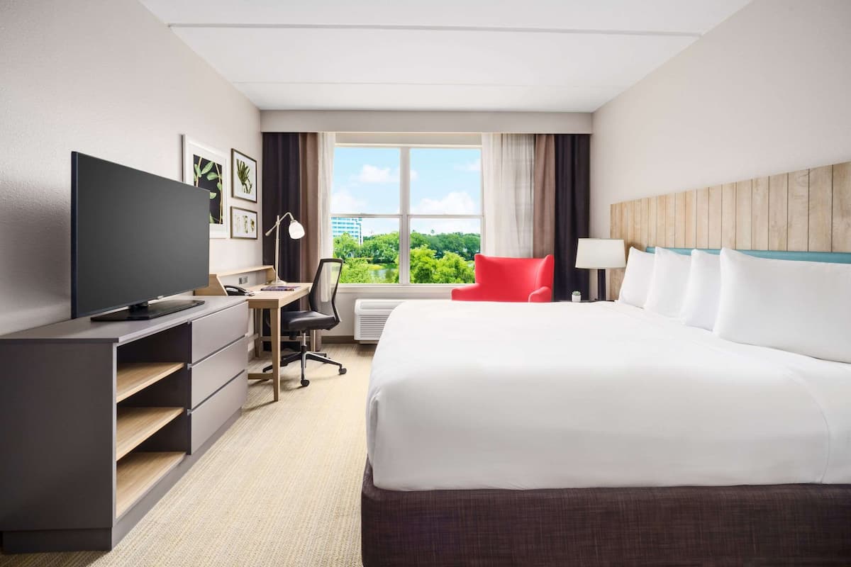 A hotel room with a large bed, a desk with a chair, a television, and a window with a view of greenery.