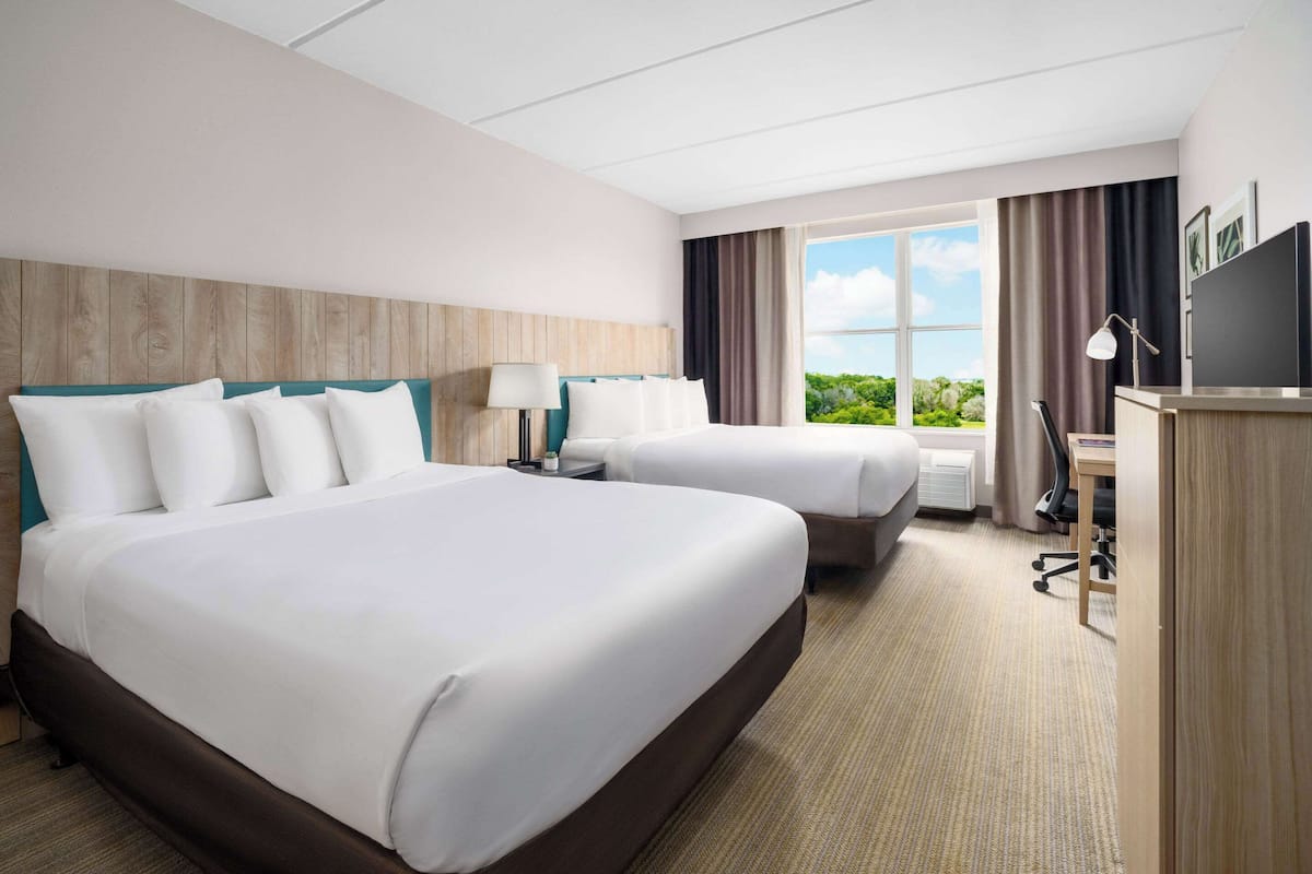 A hotel room with two beds, a desk, a chair, a TV, and a window with a view of greenery.