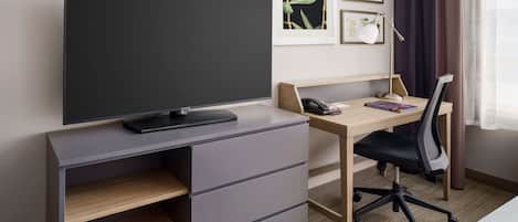 A modern hotel room with a large flat-screen TV, a wooden desk with a chair, a phone, and framed artwork on the wall.
