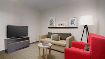 Quarto de hotel moderno com televisão de tela plana, sofá, poltrona vermelha, mesa redonda pequena e abajur.
