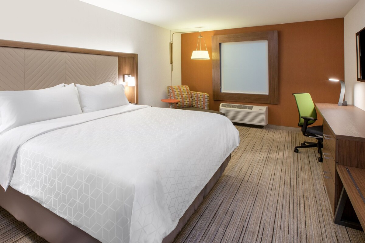 A hotel room with a large bed, a desk, a chair, a TV, and a wall-mounted heater.