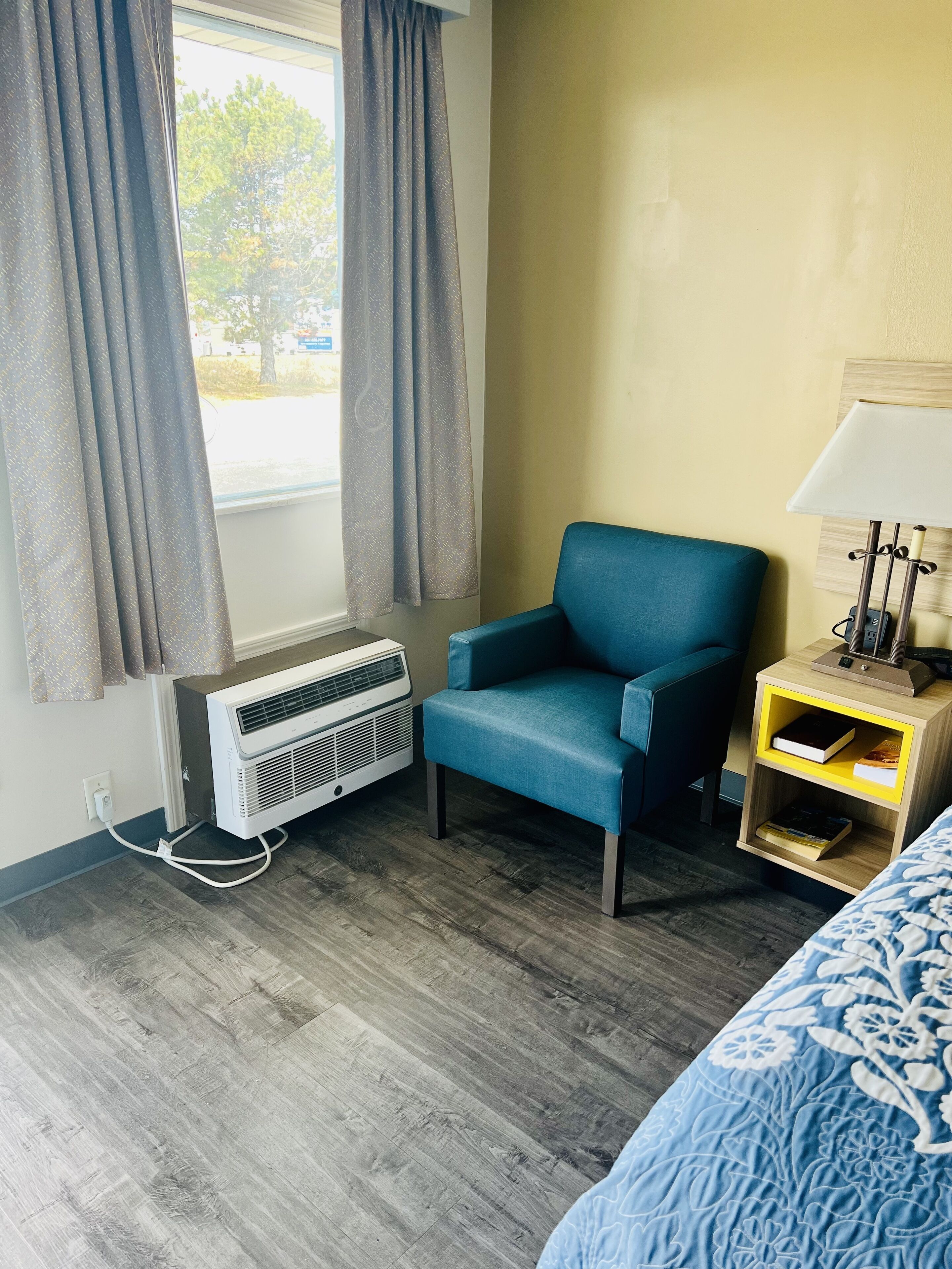 A hotel room with a blue armchair, a bedside table with a lamp, and a window with curtains.