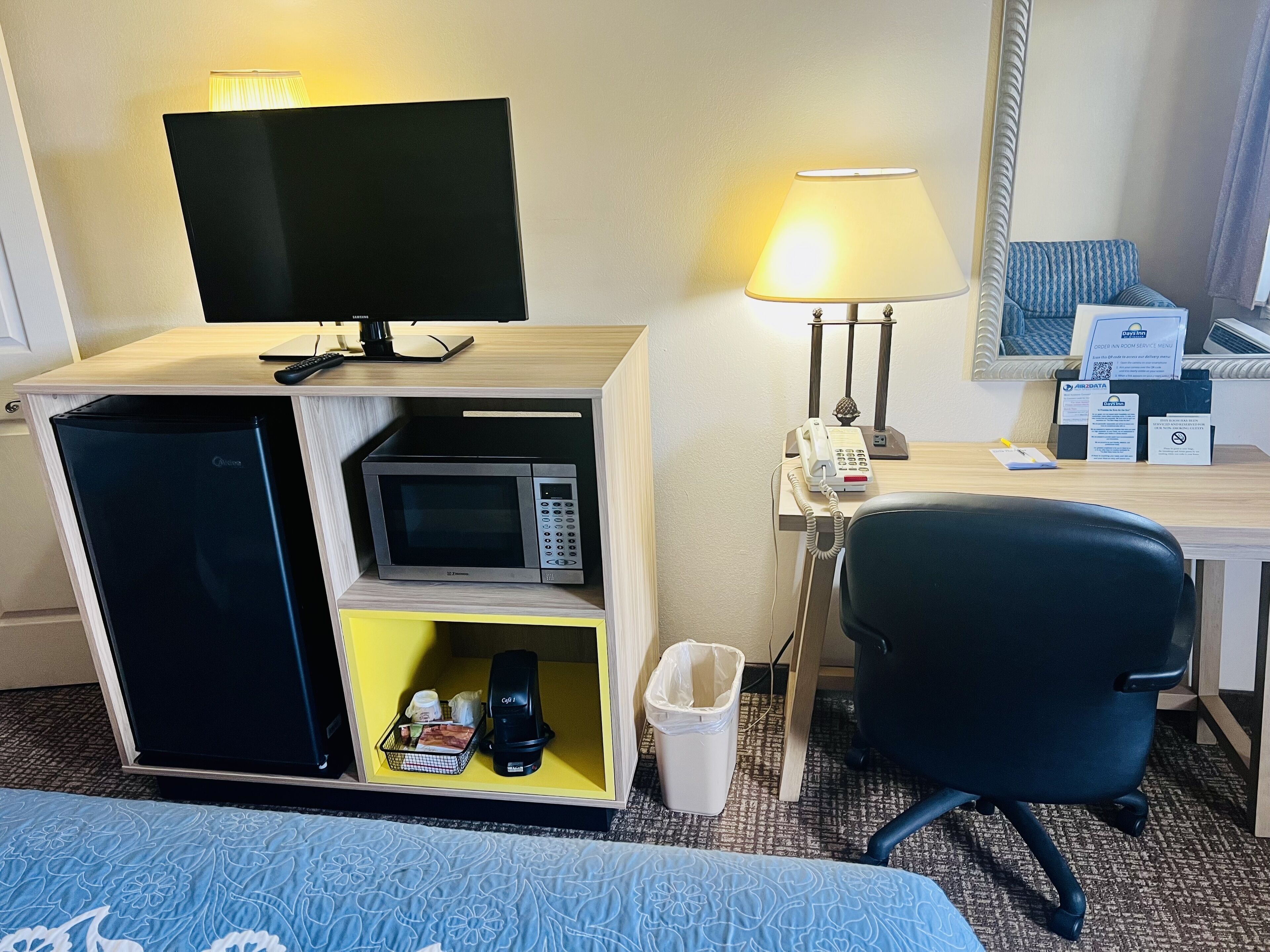 Habitación de hotel con una cama, un escritorio con lámpara, microondas, cafetera y un cubo de basura.